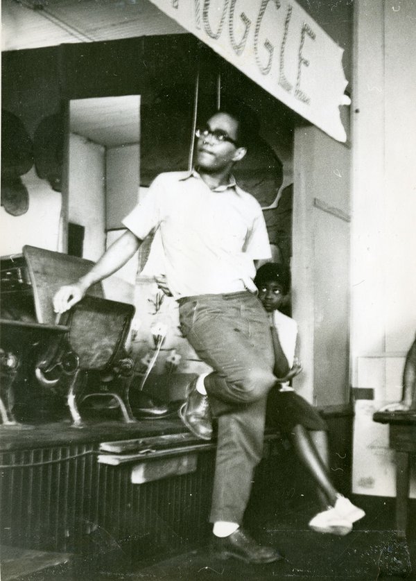 An old black & white photo of a young African American man in glasses, a white shirt and dress pants & shoes, leaning up against what appears to be chairs on a stage.