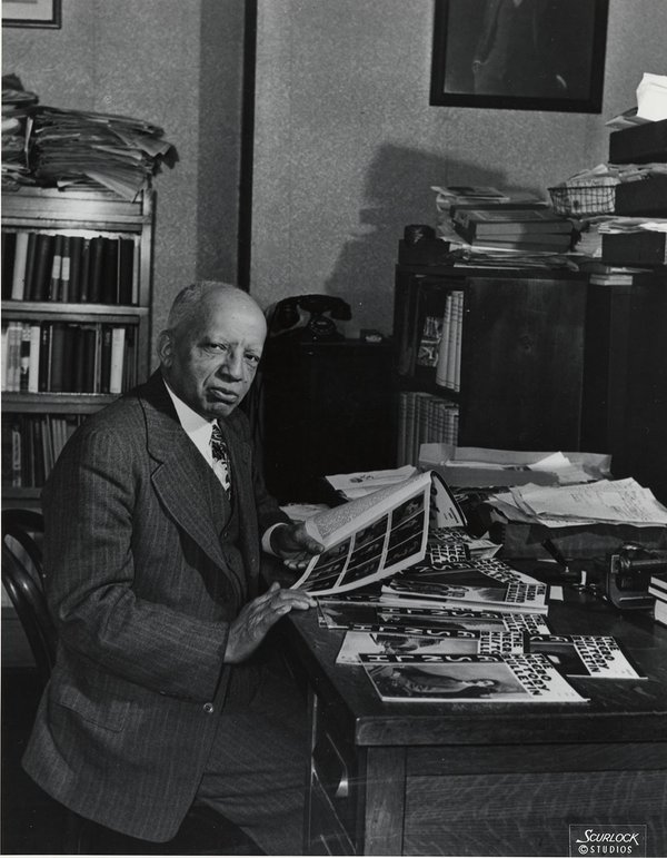 A black & white portrait of an older African American man in a suit, seated in an office in front of a desk covered in papers and magazines.