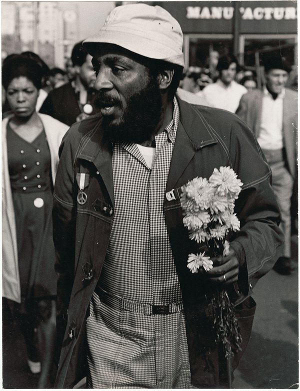 A black & white photo of a bearded African American man, walking in the midst of a crowd in a street. The man is wearing a hat, a checkered shirt & pants, and a coat with a peace medal hanging on one shoulder, and he is holding flowers in one hand.