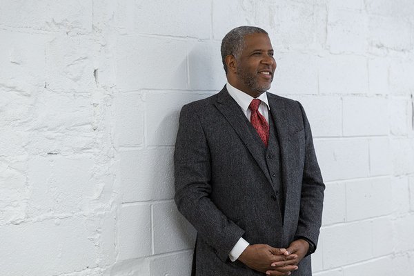 A color photo of a smiling older African American man in a dark grey suit with a white shirt and red tie, standing against a painted white concrete wall with his hands clasped in front of him.