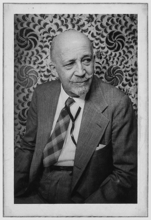 A black & white portrait of an older, bald, mustachioed, African American man in a suit sitting in front of a spiral patterned wall.