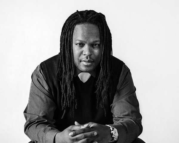 A black & white photo of an African American man with long hair braided into thin strands in a vested suit, seated and leaning forward looking at the camera, his hands clasped in front of him.