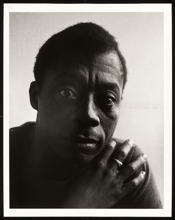 A black & white portrait of an African American man gazing at the camera/viewer, his right hand resting on his left shoulder.