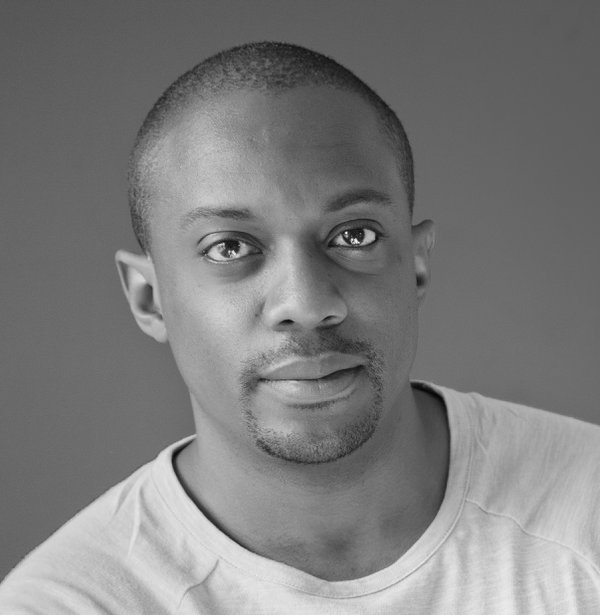 A black & white portrait of an African American man, his hair cropped back to his scalp, wearing a white t-shirt.